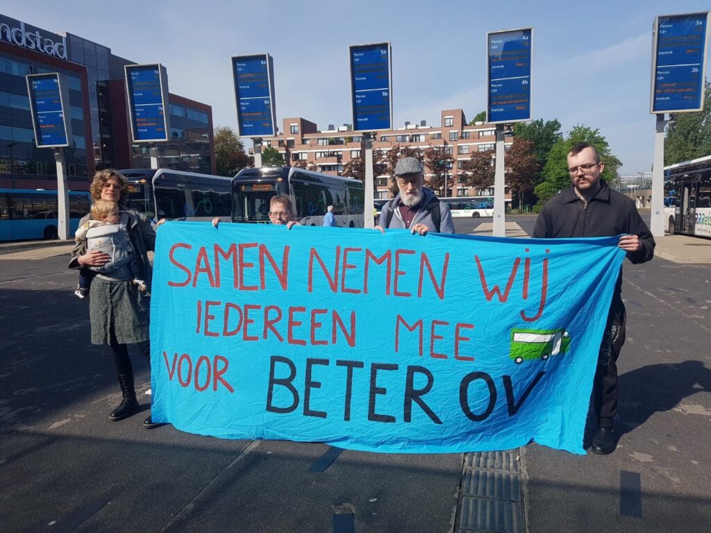 Vier mensen van de lokale groep Twente, die met een spandoek op de busremise staan. Spandoek: 'Samen nemen wij iedereen mee voor beter OV'